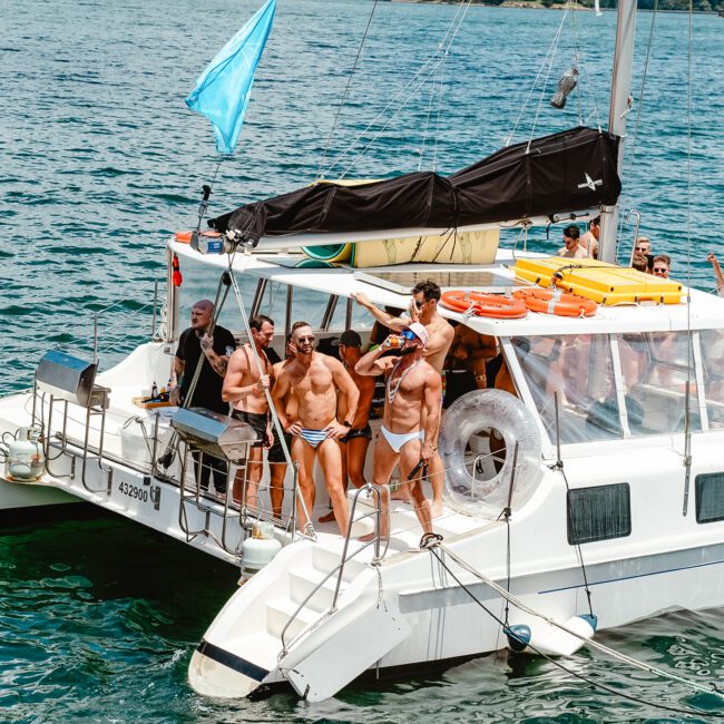 A group of people in swimsuits are gathered on a white catamaran in a sunny, blue ocean. Some stand on the deck, while others sit or lean on the railing. A light blue flag flies at the boat’s stern, and green hills are visible in the background as seagulls fly overhead.