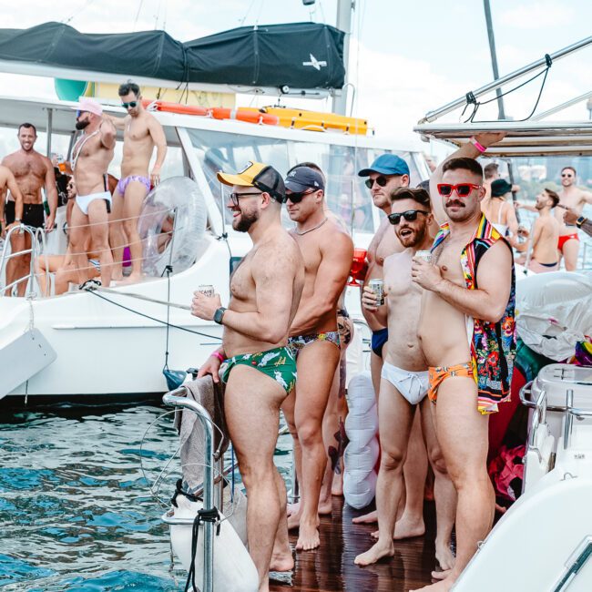 A group of men in swimwear stand on the deck of a boat, enjoying a sunny day. The boat is docked next to another vessel with more men in swimwear. They appear to be socializing and having a good time on the water, with an impressive city skyline in the background.