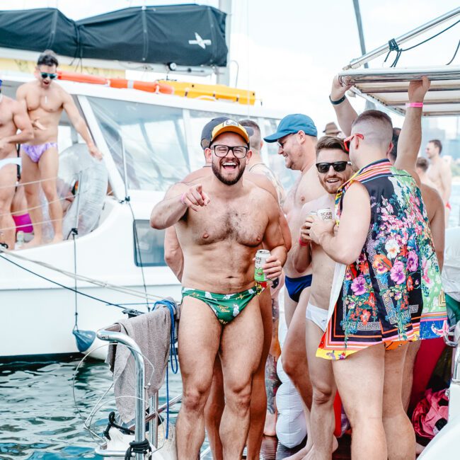 A group of people in colorful swimwear enjoy a sunny day on a boat dock. One man in green swim trunks and a yellow cap holds a drink and smiles at the camera. Others around him are chatting and laughing, creating a lively and cheerful atmosphere as children play nearby.