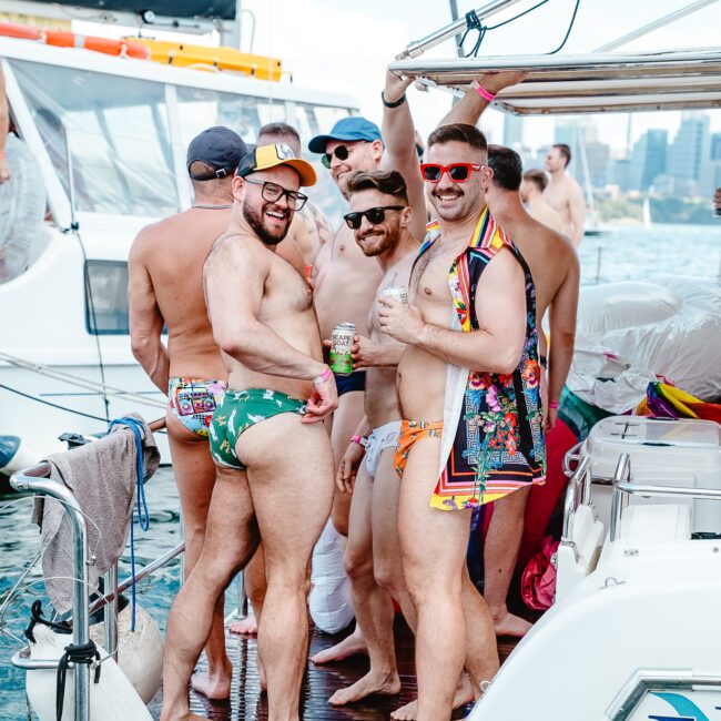 A group of men in swimwear are gathered on the deck of a boat. They are smiling and posing for the camera, with some wearing sunglasses and one in a tropical shirt. The background shows another boat and a cityscape in the distance on a sunny day, capturing the essence of their adventure.