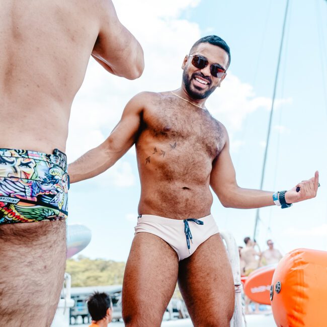 A smiling man in sunglasses and swim briefs stands on a boat, surrounded by colorful floaties and fellow participants. The background features clear skies and water, indicating summer fun on the waves.