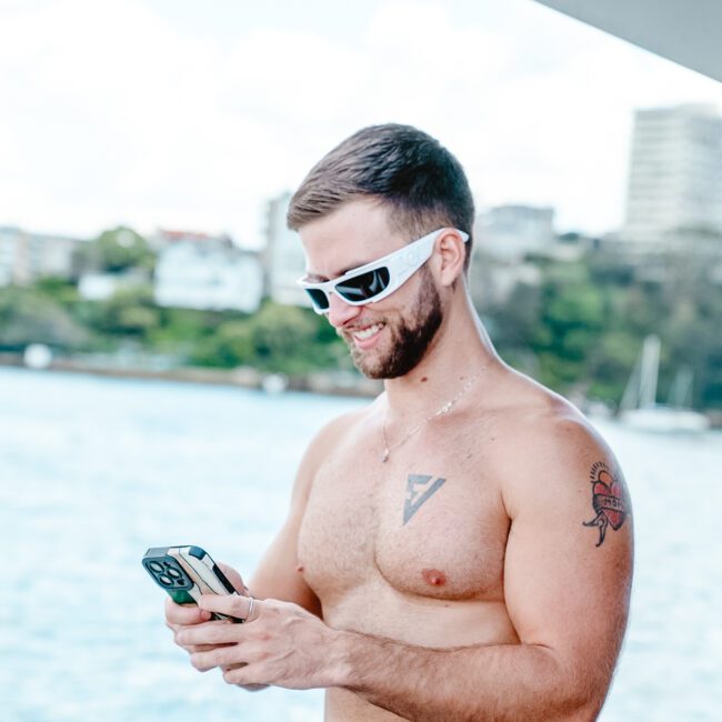 A shirtless man with sunglasses, standing on a stylish boat, glances at his phone and smiles. He has short hair, a beard, and a tattoo on his left upper arm and chest. In the background, there is water, some greenery, and buildings in the distance. The image showcases the branded logo "GAYM" at the bottom left corner.