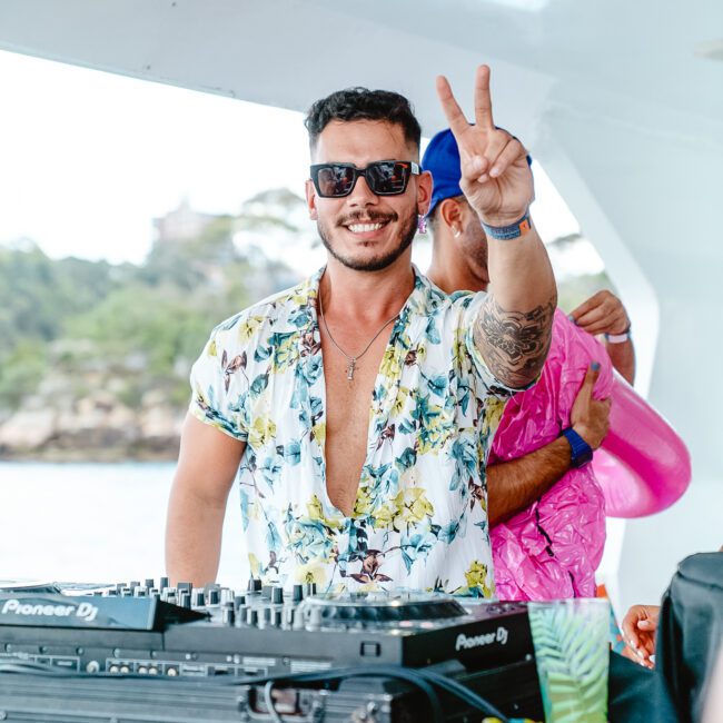 A man with a beard and sunglasses flashes a peace sign while standing behind a DJ booth on a boat party. He is wearing a partially unbuttoned floral shirt and smiling. Another person in the background, dressed in pink, is also onboard. The water and trees are visible in the distance.