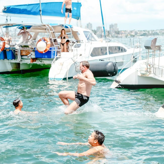 Four people swim in the sea near two docked sailboats. A man jumps into the water from one of the boats with sails gently swaying, while another person relaxes at the stern. The boats have blue canopies and flags, and the background shows a picturesque city skyline under a partly cloudy sky.