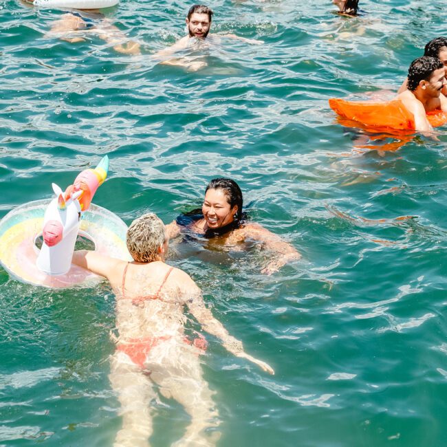 A vibrant scene of people swimming and enjoying themselves in a body of water. Floatation devices, including an inflatable unicorn ring and an orange raft, add to the fun. The atmosphere appears lively and joyful, with logos for "GAYM Entertainment" and "Gabriel Bonell" visible in the background.