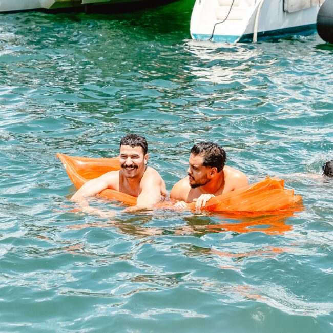 Two men are swimming in a body of water, each using an orange flotation device. They are smiling and appear to be enjoying their time. In the background, a white boat and inflatable kayak are visible.