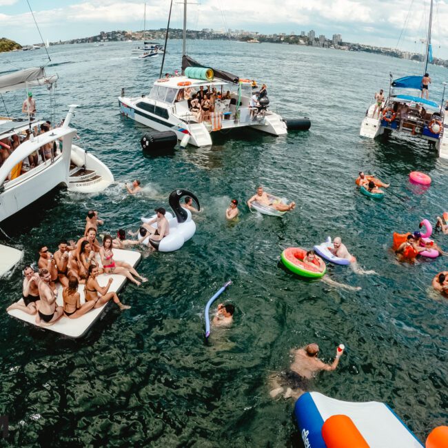 A lively scene of a boat party on a sunny day shows several people enjoying themselves on three yachts and in the water. Many float on inflatable pool toys of various shapes and colors. The Yacht Social Club Event Boat Charters hosts this celebration against the backdrop of Sydney Harbour's distant cityscape under a partly cloudy sky.