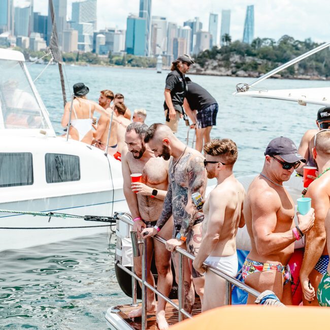 A group of men in swimwear standing and chatting on the deck of a boat, enjoying a sunny day on the water. Another boat is visible nearby, with a vibrant city skyline and skyscrapers in the background. The atmosphere appears lively and festive, enhanced by the surrounding scenic beauty.