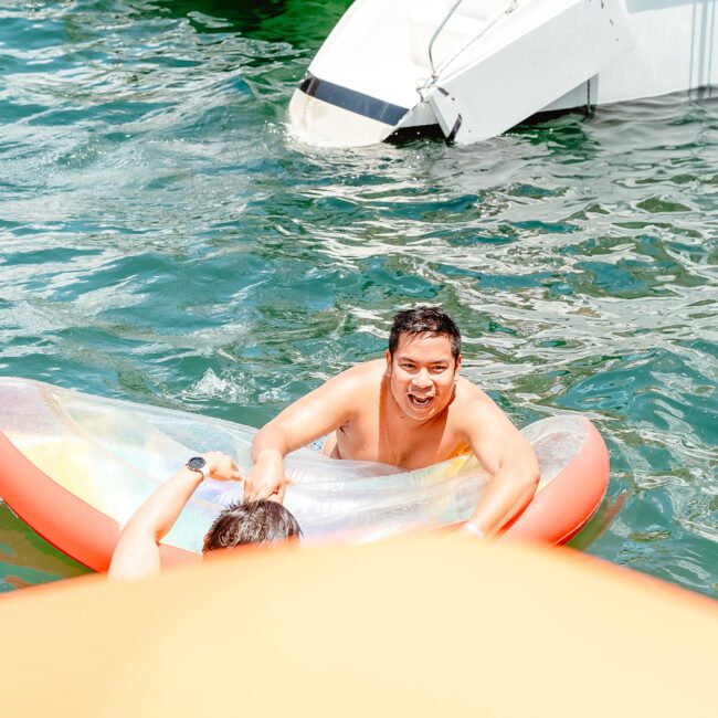 A person is smiling and having fun in the water while sitting in a transparent inflatable boat shaped like a kayak. Another person is partly visible in the water beside the boat. In the background, a sleek, white architectural structure adds to the scenic view.