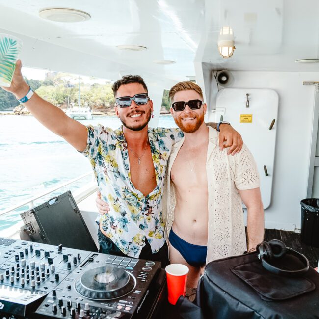 Two men on a boat enjoy a sunny day. One holds a drink aloft and wears sunglasses and a patterned shirt, while the other, with a beard, sports a sunhat, sunglasses, and an open shirt over swim trunks. DJ equipment is visible in the foreground, adding to the vibrant atmosphere of their colorful surroundings.