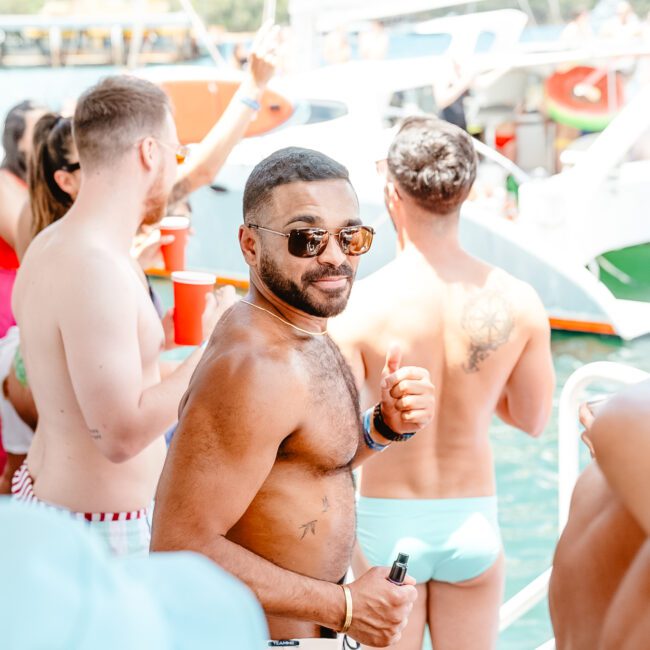 A group of people in swimwear enjoying a lively boat party. A man in sunglasses and beige shorts is shown smiling and giving a thumbs-up while holding a drink. The background features other partygoers, yachts, and a sunny, fun atmosphere.