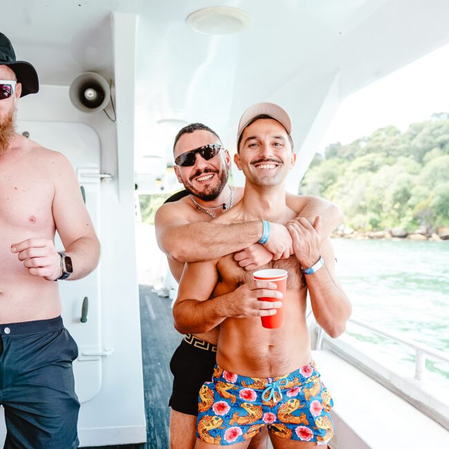 Two men are smiling and posing on a boat. One is shirtless with sunglasses, hugging the other from behind, who is holding a red cup. They are both wearing swim trunks. Another person is partially visible on the left. Trees and sparkling water create a picturesque backdrop for their joyful moment.