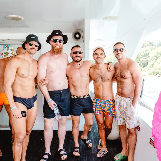 A group of five men, all shirtless, stand and smile on a boat in their colorful swim trunks and hats. In the background, one man is partially visible, also in swimwear. The boat is on a serene body of water with lush greenery visible onshore.