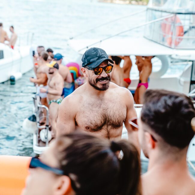 A group of people enjoying a boat party on a sunny day. The focus is on a shirtless man, wearing sunglasses and a cap, smiling and interacting with others. Vibrant boats and more partygoers can be seen in the background on the water, adding to the lively atmosphere.