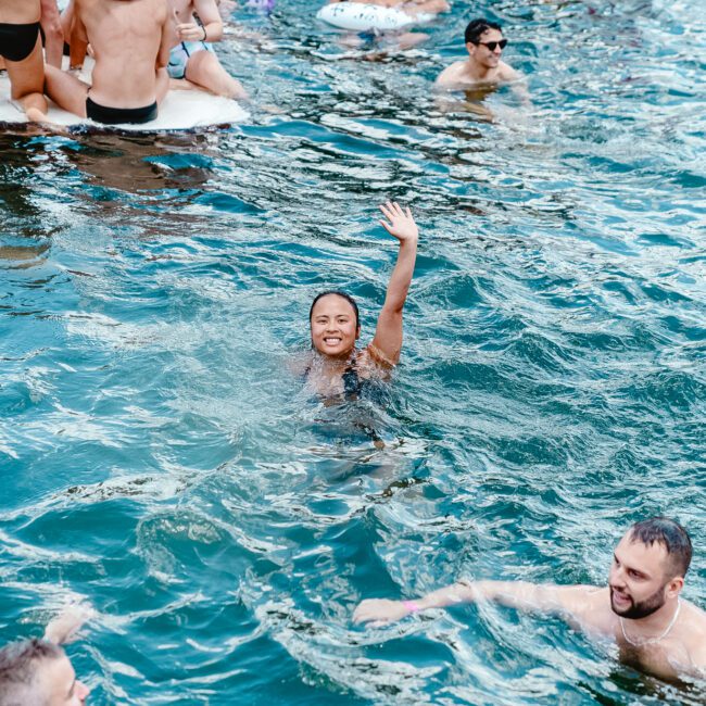 A group of friends enjoying a swim in a pool. In the center, a person is smiling and waving while swimming. Others are scattered around either floating, standing, or playing water games. The water is clear and vibrant, creating an inviting atmosphere.