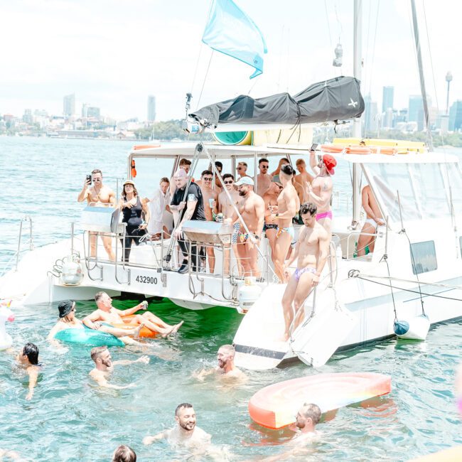 A group of people enjoy a sunny day on a boat and in the water. The boat is anchored, with people standing and sitting on it, some wearing swimsuits and sunglasses. In the water, others float on colorful inflatable toys. A stunning city skyline is visible in the background.