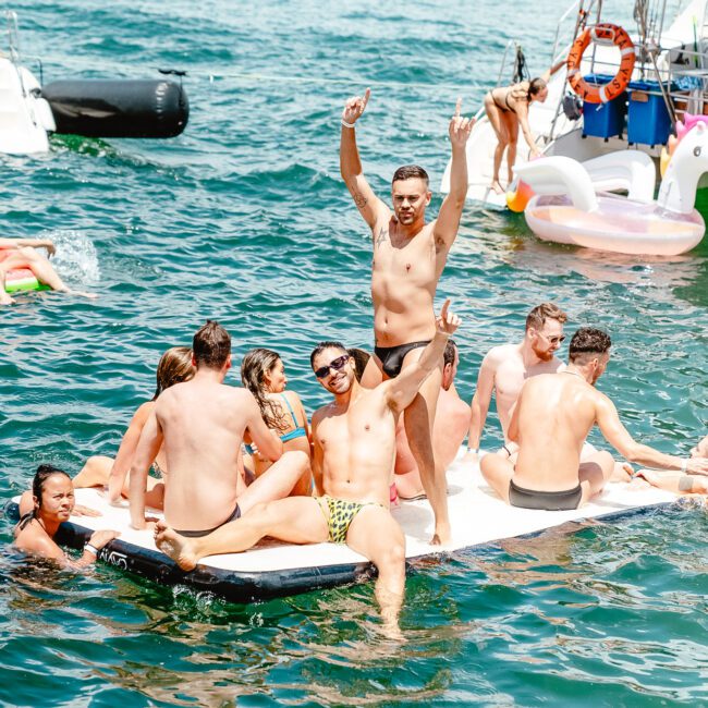 A group of people are floating on a rectangular paddleboard in a sunny body of water. One man standing raises his arms joyfully, while others are seated or lounging. Inflatable toys, a kayak, and another boat are visible in the scenic background.