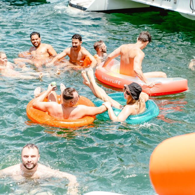 A group of people are enjoying a sunny day in the water, with some in inflatable rings and others swimming or relaxing. They appear to be socializing and having a good time, surrounded by boats. The atmosphere is lively and cheerful, with music playing from nearby yachts.