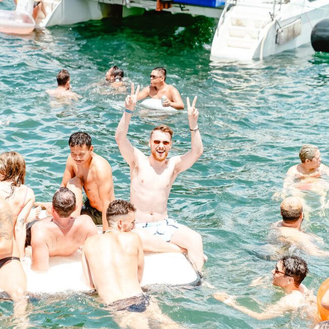 People are enjoying a sunny day in the water near a boat. One person in the center is sitting on an inflatable raft, smiling, and making peace signs with both hands. Others are swimming, relaxing on paddleboards and inflatables, and conversing. The scene is bright and lively.