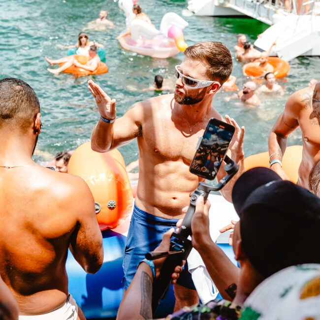 A group of people enjoy an outdoor pool party on a sunny day. The men in the foreground are shirtless, wearing sunglasses and shorts, and gesturing animatedly. Others in the background are lounging on floaties in the water, while nearby, kayaks glide across the serene lake.