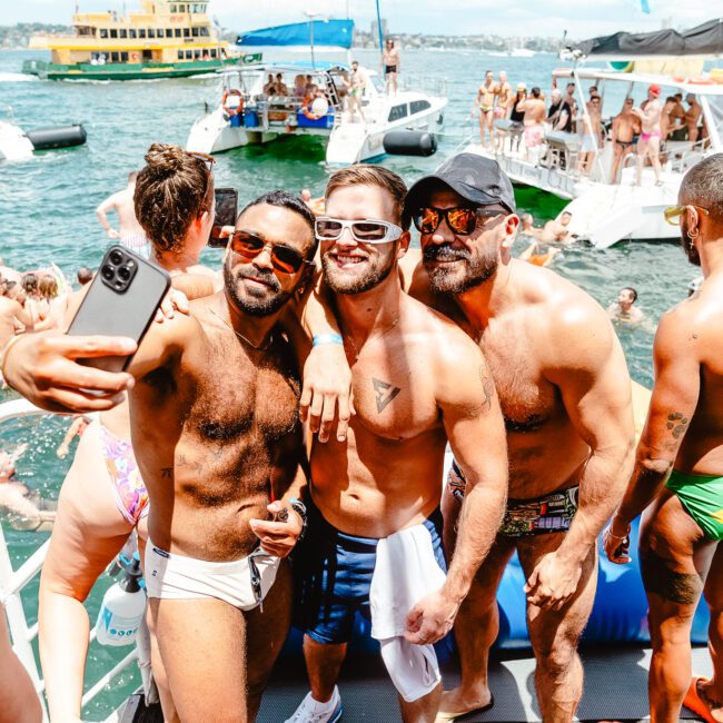 A group of four men in swimwear take a selfie on a boat during a sunny day. They are surrounded by other people, some on a nearby yacht, enjoying a lively summer party on the water. The background features more boats and a sparkling body of water.