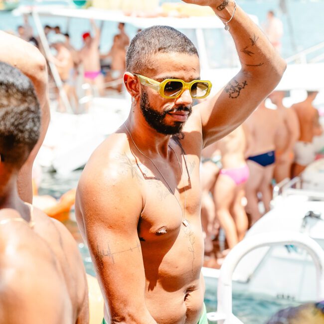 A shirtless man wearing yellow sunglasses and green swim trunks dances on a boat. He has a beard, various tattoos, and a raised arm. Several other people in colorful swimwear are present on the boat and in the background, enjoying what seems to be a lively, sun-soaked outdoor party.