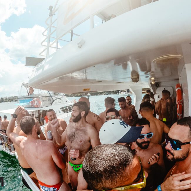 A group of people are enjoying a lively party on a boat in sunny weather. Many are wearing sunglasses and swimwear, and some are dancing to upbeat music. Another boat is cruising nearby. The atmosphere appears festive and social, with trees and a bridge in the background.