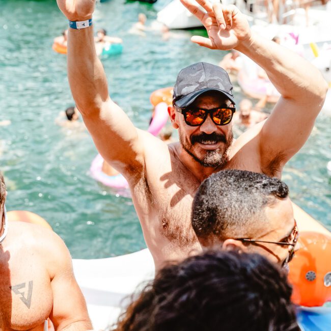 A man wearing sunglasses and a cap raises his arms in celebration at a lively outdoor pool party. Shirtless and smiling, he is surrounded by other joyous partygoers in swimwear. In the background, people are enjoying themselves in the water with inflatables and kayaks.