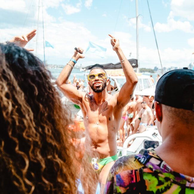 A shirtless man wearing sunglasses dances exuberantly with his arms raised in the middle of a crowded boat party. Other partygoers groove around him, with boats and a bright sky in the background. The festive atmosphere is lively and vivacious.