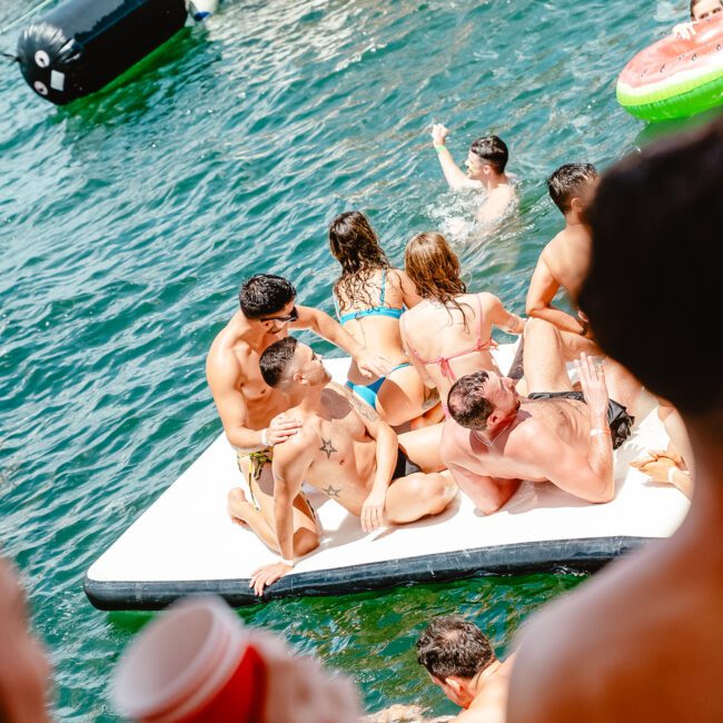 A lively group of people in swimwear enjoying a float party on a lake surrounded by boats. Some are sitting, while others splash in the water. One person in the foreground holds a red cup. Inflatable toys and pool floats add to the fun. Logos for "GAYM" and "Yacht Social Club" are visible in the corners.