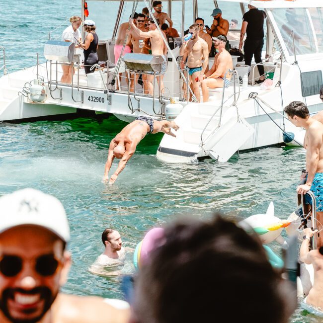 A lively group in swimwear enjoys a sunny day on and in the water near a catamaran. One person is mid-dive while others watch or swim in the clear blue water. Logos for GAYM and "Côte Soleil" are visible, enhancing the vibrant atmosphere.