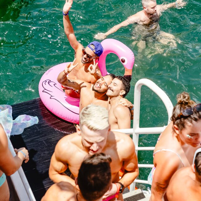 A group of people enjoying a pool party on a sunny day. Several individuals are in the water, with three friends posing on a pink flamingo float. Others are standing by a ladder leading into the pool, with colorful inflatables and pool toys visible in the water. It’s an idyllic summer scene filled with fun and laughter.