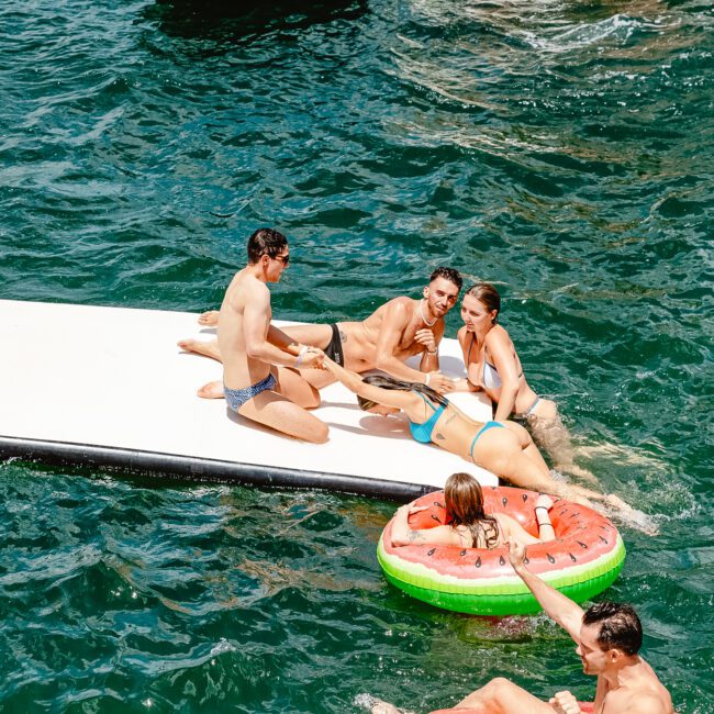 A group of young adults are enjoying a sunny day on the water. Some are lounging on a floating platform, while others are swimming or relaxing on colorful inflatable pool toys. The deep green water sparkles under the cheerful atmosphere.