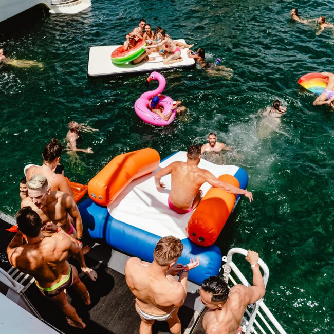 A group of people enjoying a sunny day on a boat and in the water. Some are on inflatable floats, including a pink flamingo and rainbow-colored designs. Others are swimming or socializing on the boat deck, with some standing, sitting, and lying down. The mood is all about fun and relaxation.