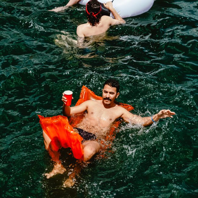 A man lounges on an orange float in the water, holding a red cup and smiling. The water around him is dark green. Another person is partially visible in the background, enjoying the day on a white inflatable.