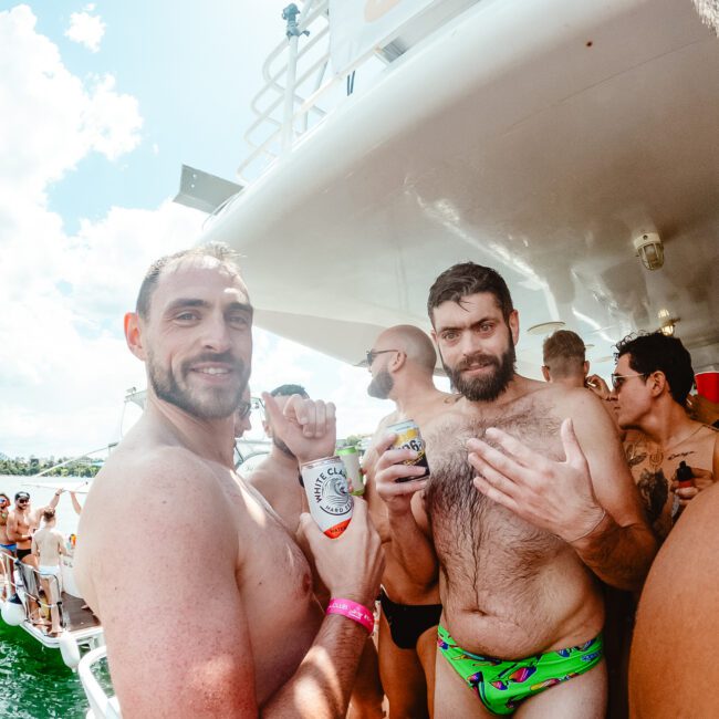 Two men in swimwear stand on a boat holding cans of drink, smiling at the camera. They are surrounded by other people, enjoying a sunny day out on the water with a festive vibe. The atmosphere is lively, with logos "GAYM" and "Upstart Social Club" visible in the corners.