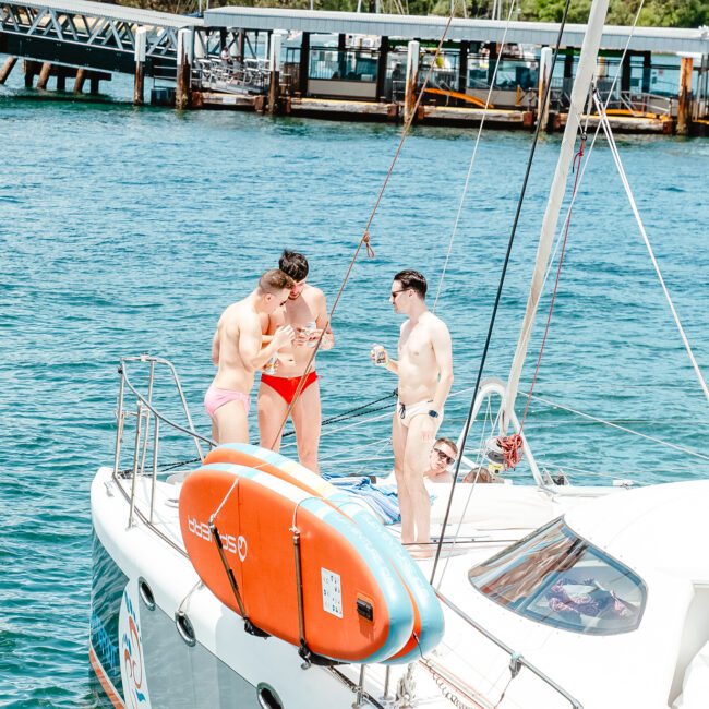 Three people on a sailboat are enjoying a sunny day on the water. Two are standing and conversing, while the third, in a red swimsuit, is looking down at the lifebuoy. The boat is anchored near a pier, with other equipment visible on deck.