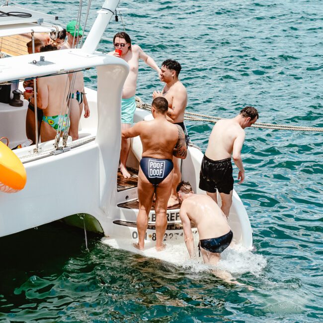 A group of people in swimwear are on the back of a boat, with one person stepping into the water. A life preserver is attached to the boat. The clear blue sea surrounds them, and some people are holding drinks, enjoying a refreshing escape on the water.