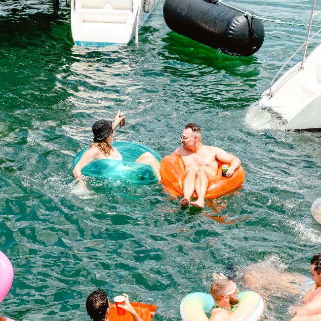 People are floating on inflatable pool toys in a body of water next to a boat. One woman in a green floatie and a man in an orange floatie are interacting, while others drift nearby. The atmosphere is relaxed and festive, suggesting an enjoyable leisure activity on a sunny day, possibly during summer holidays.