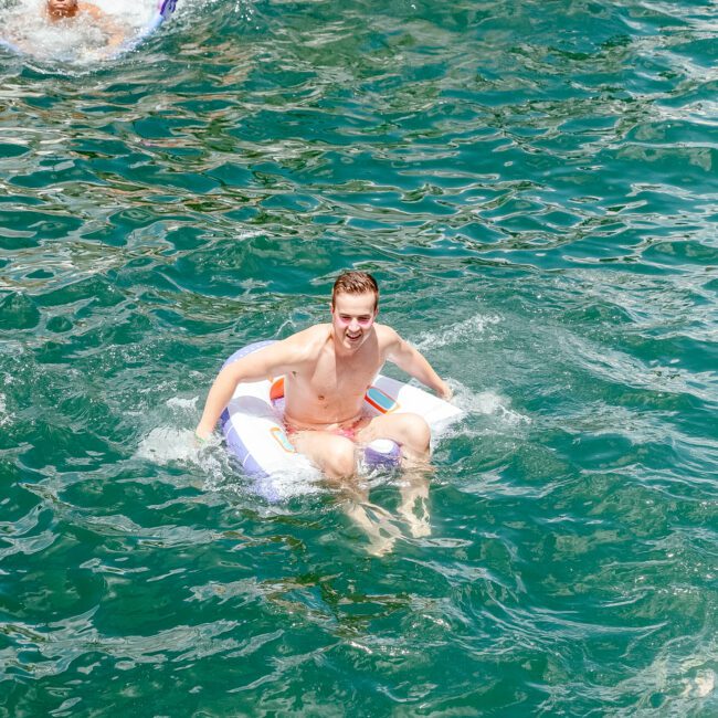 A man floats on an inflatable pool lounger in a body of water with a smile, while another person swims nearby. In the background, more people enjoy the dock. Water splashes around them, creating a fun and lively summer scene. Branding logos are subtly visible in the corners.