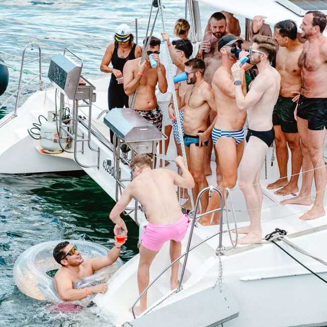 A group of people on a boat are enjoying drinks and socializing. One person is in the water, holding a drink and interacting with those on board. Most are wearing swimsuits, and the scene appears festive and lively. The boat is anchored in calm, sunlit waters.