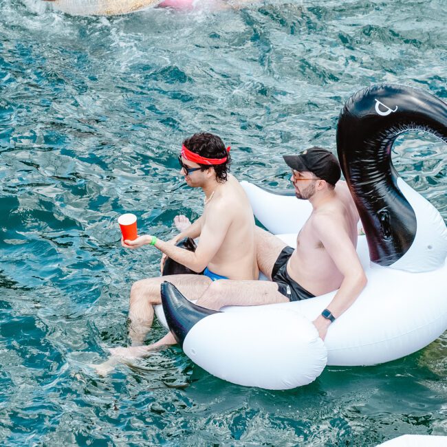 Three men float in a pool on inflatable toys. One man sits on a large white swan float, holding a red cup, while another sits behind him enjoying the calm. The third man relaxes on an ocean-themed blue float. The background shows clear water and colorful inflatable toys.