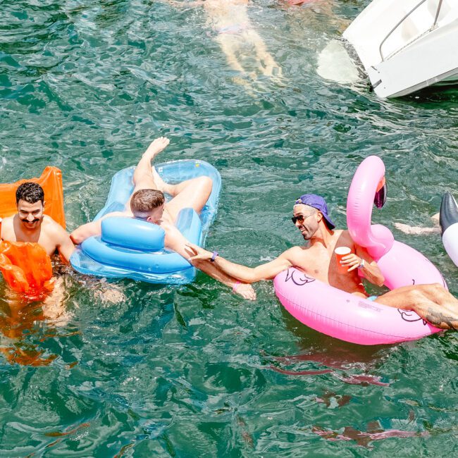 A group of people enjoy a sunny day swimming and floating on various inflatable items in a body of water. One person in a pink flamingo float holds hands with another person on a blue unicorn float, creating a colorful scene. A white slide from a nearby structure leads into the water.