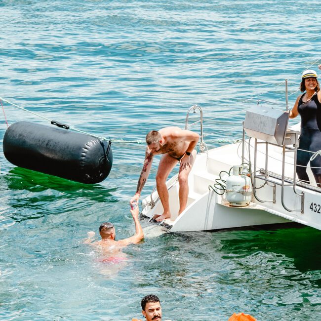 A group of people enjoy a sunny day on a yacht in the water. One person is swimming, another is on the yacht helping them up. Others are waving and smiling. Another boat is docked nearby, and the background shows clear blue water.