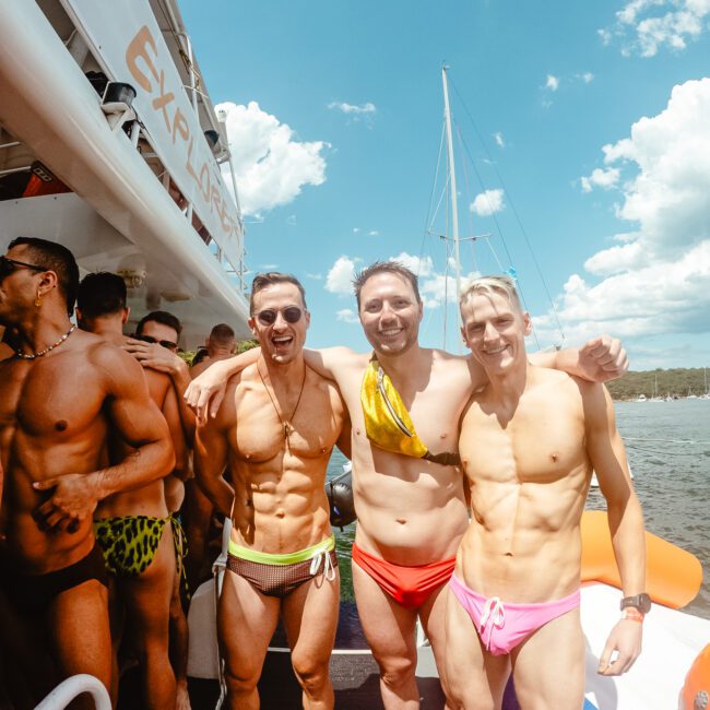 Three smiling men in colorful swimwear pose for a photo on a boat during a sunny day, surrounded by others enjoying the lively event. Clear skies and sparkling water create a perfect backdrop. Logos indicating "GAYM" and "Uptown Social" are visible in the corners of the vibrant scene.