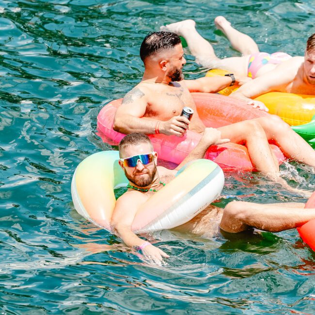 Two people floating in a body of water on inflatable rings. One, closer to the camera, sports a colorful unicorn float, dons sunglasses and holds a drink. The other person relaxes further back on a simple round ring. Logos for GAYM and Uptown Social appear at the bottom corners.