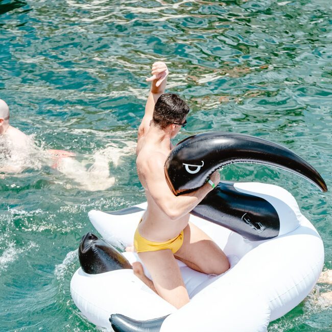 A person in a yellow swimsuit rides a large, inflatable orca in a sparkling blue body of water, raising one arm triumphantly. Another swimmer is in the background, gliding through the water. The scene is bright and sunny and feels full of summer joy.