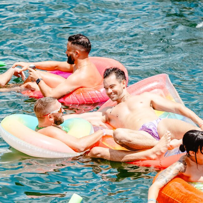 A group of friends enjoying a sunny day at the lake, floating on colorful inflatables. They appear to be laughing and chatting, with some splashing water. The atmosphere is festive and lively, with the vibrant hues of the unicorn-shaped inflatables contrasting beautifully with the blue water.