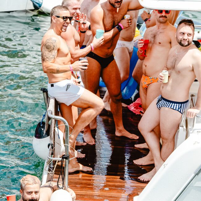 A group of men are on a boat, enjoying drinks and socializing. They are casually dressed in swimwear, some with hats and sunglasses. The atmosphere is lively and relaxed, surrounded by sparkling water with other boats in the background, making for a perfect day on the lake.