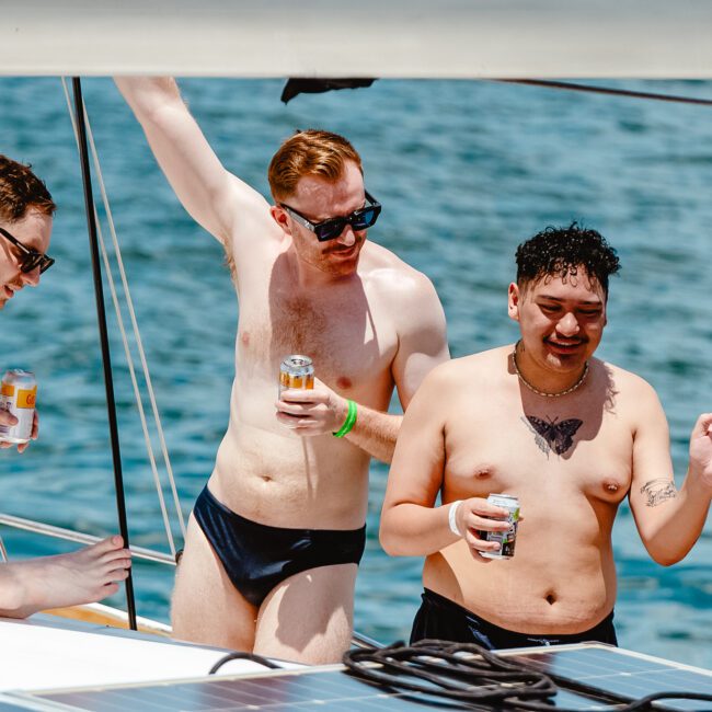 Four men in swimwear are on a boat, enjoying a sunny day on the water. Some are holding canned beverages and are engaged in conversation and laughter under the clear blue sky. The picturesque sea is visible in the background, creating a relaxed and joyful atmosphere.